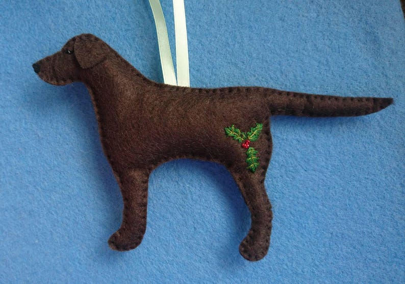 May include: A brown felt dog ornament with a holly sprig embroidered on its side. The ornament is hanging from a white ribbon.