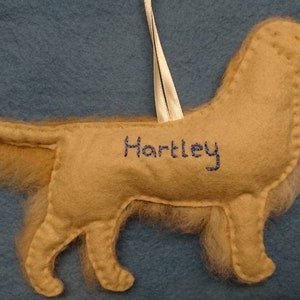 K&ouml;nnte beinhalten: Ein beigefarbener Filzhund-Anh&auml;nger mit einem wei&szlig;en Band und dem Namen "Hartley", der auf die Seite gestickt ist.