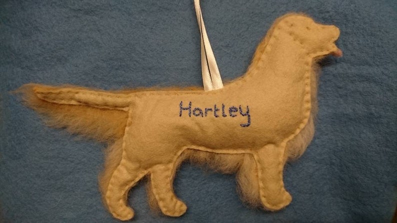 K&ouml;nnte beinhalten: Ein beigefarbener Filzhund-Anh&auml;nger mit einem wei&szlig;en Band und dem Namen "Hartley", der auf die Seite gestickt ist.