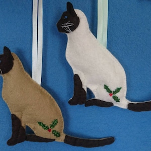May include: Two felt Siamese cat ornaments with brown and white markings and a hand embroidery green holly leaf design with three red glass beads berries. The cats are sitting with their tails stretched straight out behind their bbodies. One cat is fawn and chocolate, the other is white and chocolate coloured.