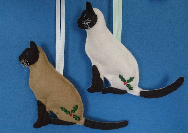 May include: Two felt Siamese cat ornaments with brown and white markings and a hand embroidery green holly leaf design with three red glass beads berries. The cats are sitting with their tails stretched straight out behind their bbodies. One cat is fawn and chocolate, the other is white and chocolate coloured.