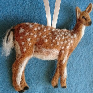 May include: A brown and white needle felted deer ornament with a white tail. The deer has a white ribbon attached to its back for hanging.