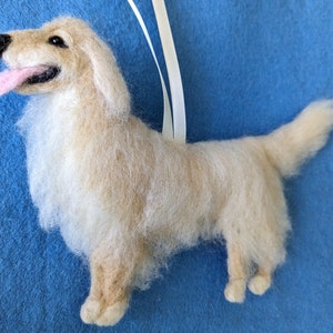 K&ouml;nnte beinhalten: Ein cremefarbener Filz-Anh&auml;nger eines Golden Retriever Hundes mit herausgestreckter rosa Zunge. Der Hund steht auf allen vieren und hat ein wei&szlig;es Band an seinem R&uuml;cken.