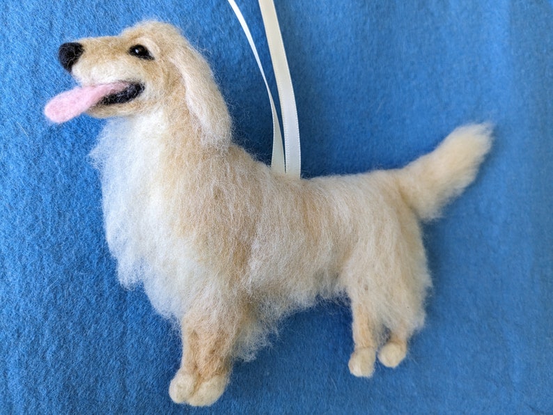 K&ouml;nnte beinhalten: Ein cremefarbener Filz-Anh&auml;nger eines Golden Retriever Hundes mit herausgestreckter rosa Zunge. Der Hund steht auf allen vieren und hat ein wei&szlig;es Band an seinem R&uuml;cken.