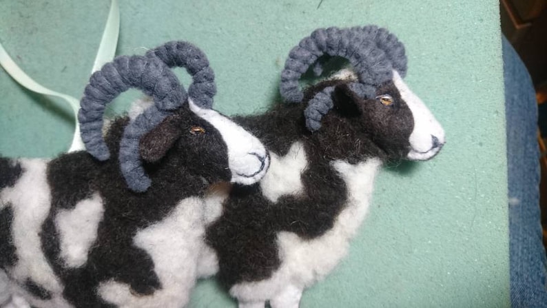 Puede incluir: Dos ovejas de fieltro blanco y negro con cuernos grises. Las ovejas est&aacute;n tumbadas con la cabeza girada hacia un lado.
