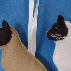 May include: Two felt Siamese cat ornaments, one brown and one white, with blue eyes. Both cats are facing to the right and have a white ribbon attached to the top.