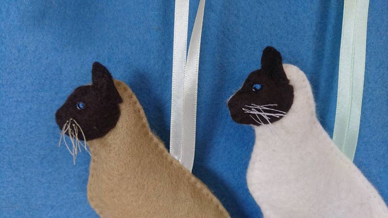 May include: Two felt Siamese cat ornaments, one brown and one white, with blue eyes. Both cats are facing to the right and have a white ribbon attached to the top.