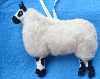 May include: A white wool felt sheep ornament with black markings on its face and legs. The sheep is standing on a blue background.