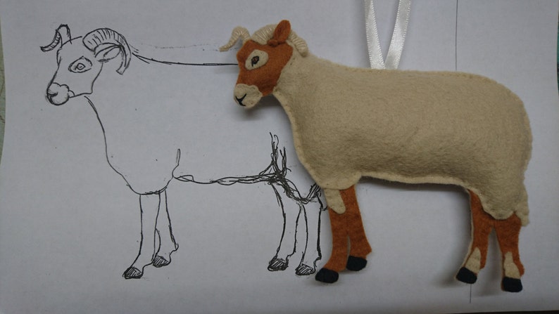 May include: A beige felt sheep ornament with brown legs and a black nose. The sheep is hanging from a white ribbon. A line drawing of a sheep is behind the ornament.