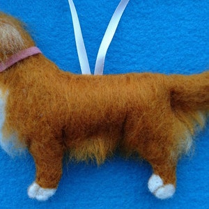 May include: A brown and white felt dog ornament with a pink collar. The dog is standing on a blue surface and has a white ribbon attached to its back.