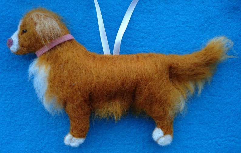 May include: A brown and white felt dog ornament with a pink collar. The dog is standing on a blue surface and has a white ribbon attached to its back.