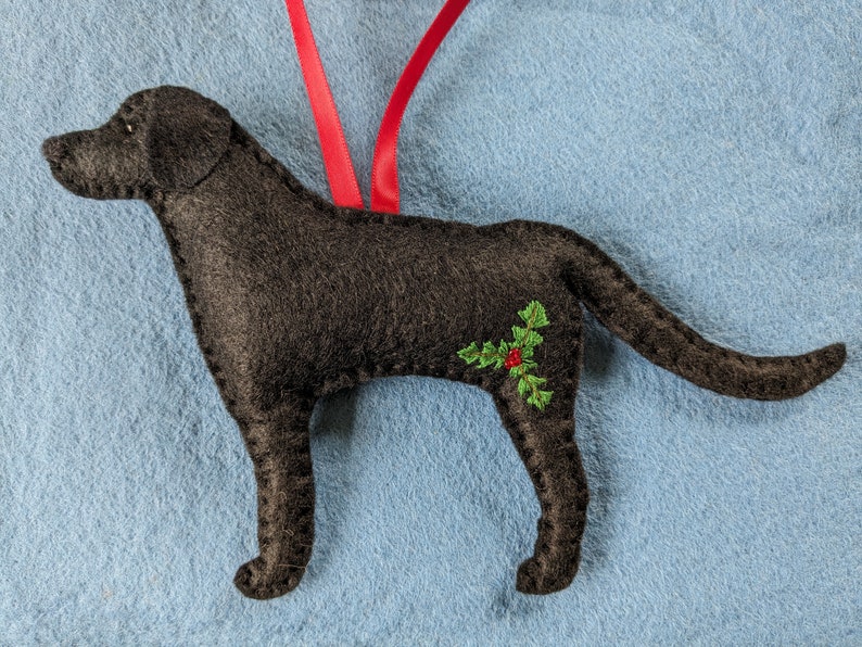 May include: Black felt ornament of a Labrador Retriever dog with green holly and red berries embroidered on its side.