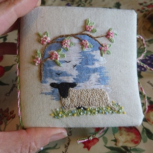 May include: A white fabric square with a hand-stitched embroidery design of a sheep under a blooming tree. The tree has pink flowers and green leaves. The sheep is white with a black face and is standing in a field of green grass with yellow flowers. The fabric has a pink and green border.