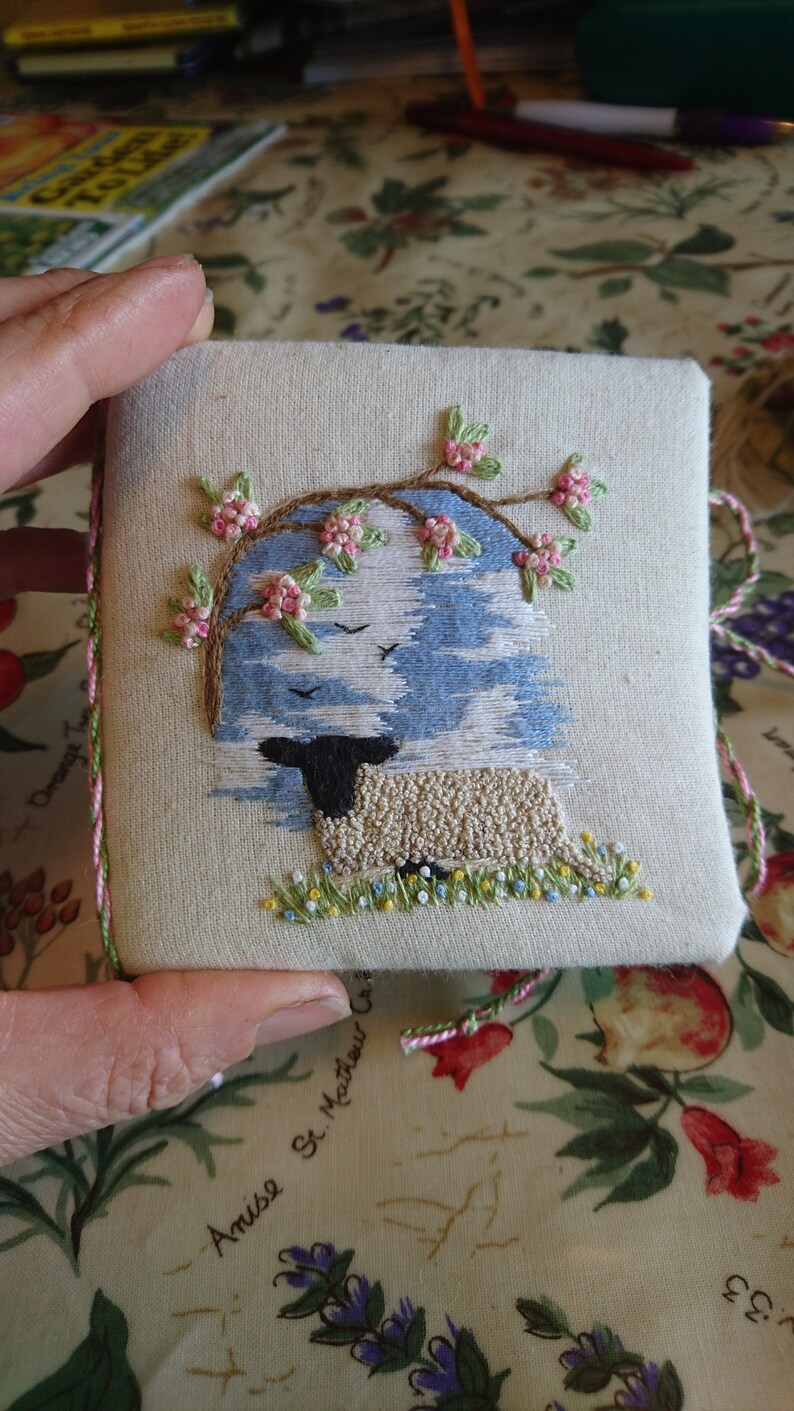 May include: A white fabric square with a hand-stitched embroidery design of a sheep under a blooming tree. The tree has pink flowers and green leaves. The sheep is white with a black face and is standing in a field of green grass with yellow flowers. The fabric has a pink and green border.
