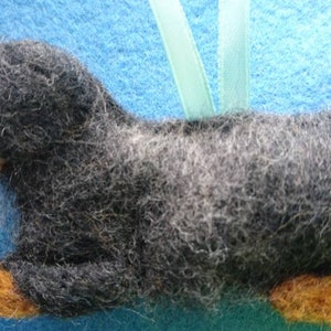 May include: A needle-felted dog ornament featuring a long body covered in dark grey and black wool fibres. The dog has a tan-coloured muzzle and paws, a small black nose, and a dark eye. A light green ribbon loop is attached to the top for hanging.