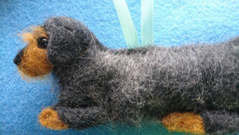 May include: A needle-felted dog ornament featuring a long body covered in dark grey and black wool fibres. The dog has a tan-coloured muzzle and paws, a small black nose, and a dark eye. A light green ribbon loop is attached to the top for hanging.