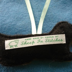 May include: A black and brown felt dog ornament with a light green ribbon loop for hanging. A white fabric label is stitched to the centre, featuring a green sheep icon and the text: HAND MADE BY PAULA AT Sheep In Stitches.