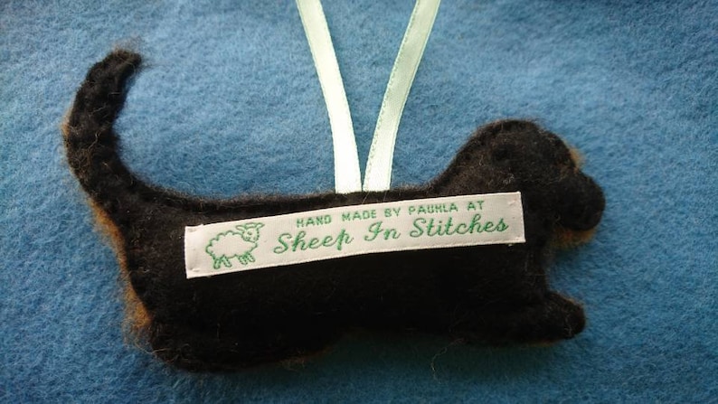 May include: A black and brown felt dog ornament with a light green ribbon loop for hanging. A white fabric label is stitched to the centre, featuring a green sheep icon and the text: HAND MADE BY PAULA AT Sheep In Stitches.