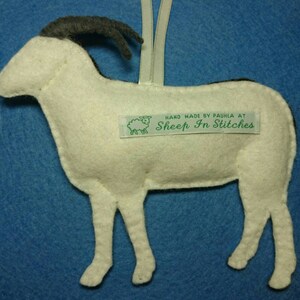 Puede incluir: Adorno de oveja de fieltro blanco con cuernos de fieltro negro. El adorno tiene una etiqueta de fieltro verde que dice "Hand Made by Paula at Sheep In Stitches".