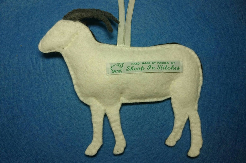 Puede incluir: Adorno de oveja de fieltro blanco con cuernos de fieltro negro. El adorno tiene una etiqueta de fieltro verde que dice "Hand Made by Paula at Sheep In Stitches".
