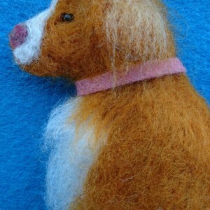 May include: A brown and white needle felted dog with a pink collar. The dog is looking to the left.