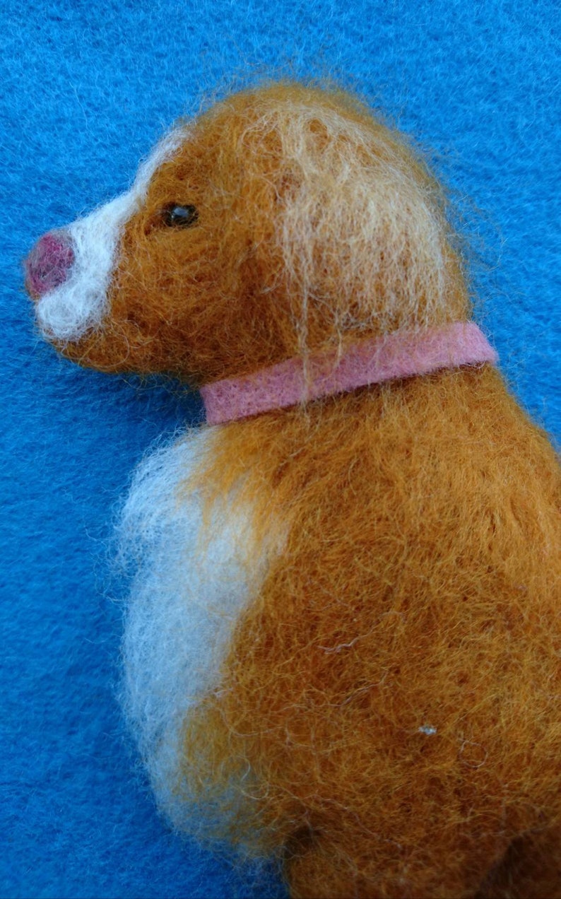 May include: A brown and white needle felted dog with a pink collar. The dog is looking to the left.