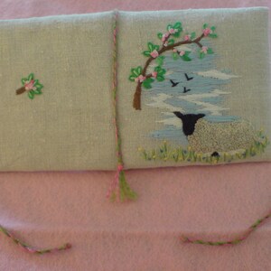 May include: A light blue linen pouch with a green and pink embroidered design of a sheep and a tree with pink flowers. The pouch has a green and pink tassel.