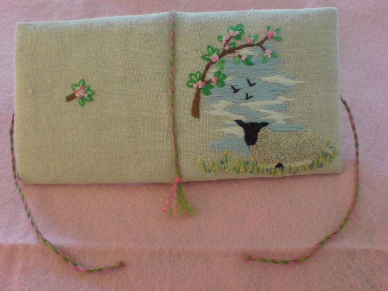 May include: A light blue linen pouch with a green and pink embroidered design of a sheep and a tree with pink flowers. The pouch has a green and pink tassel.