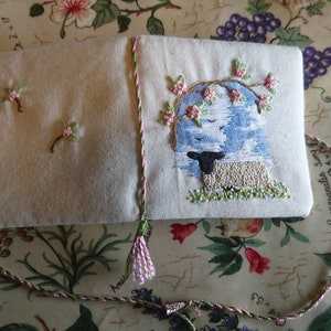 May include: A small, white fabric book with a pink tassel. The front cover features an embroidered design of a black and white sheep standing under a tree with pink flowers. The text "Comegranates song" is embroidered on the back cover.