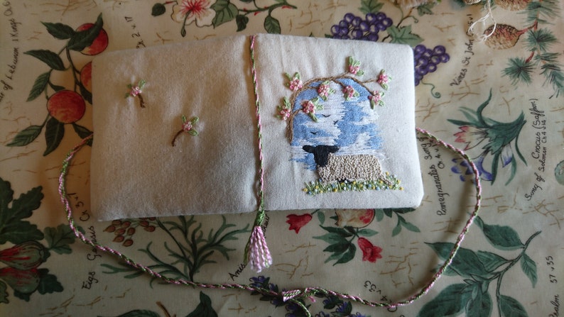 May include: A small, white fabric book with a pink tassel. The front cover features an embroidered design of a black and white sheep standing under a tree with pink flowers. The text "Comegranates song" is embroidered on the back cover.