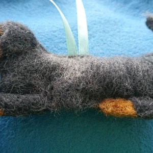 May include: A needle-felted dog ornament featuring a charcoal grey body with tan accents on the muzzle, paws, and tail tip. The figure has a textured, fuzzy appearance and includes a light green ribbon loop attached to the back for hanging.