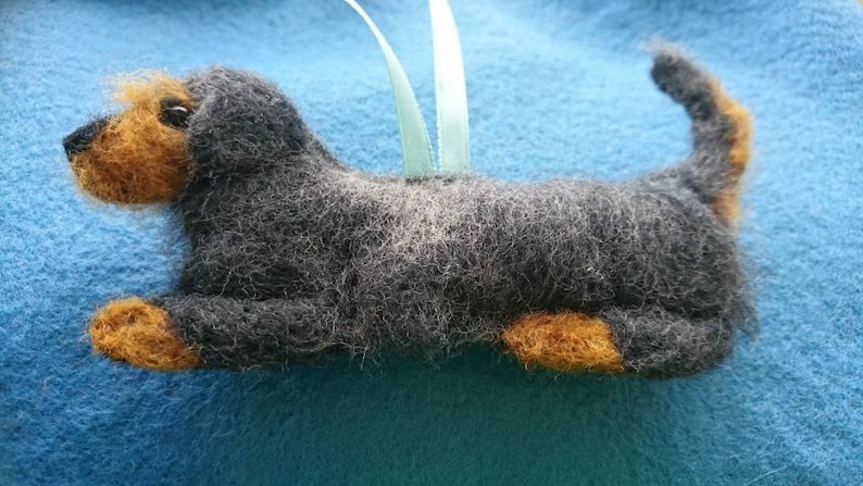 May include: A needle-felted dog ornament featuring a charcoal grey body with tan accents on the muzzle, paws, and tail tip. The figure has a textured, fuzzy appearance and includes a light green ribbon loop attached to the back for hanging.