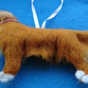 May include: A hand-felted, brown and white dog ornament with a pink collar. The dog is standing on a blue surface.