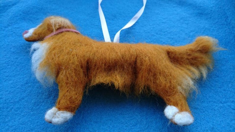 May include: A hand-felted, brown and white dog ornament with a pink collar. The dog is standing on a blue surface.