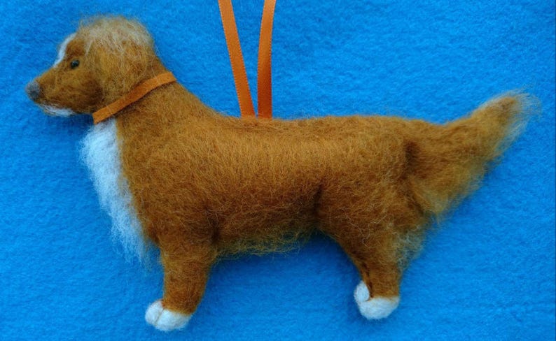 May include: A hand-felted brown and white dog ornament with a white chest and a fluffy tail. The dog is wearing an orange felt collar.