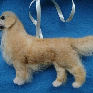 K&ouml;nnte beinhalten: Ein handgefilzter Golden Retriever Hundefiguren-Anh&auml;nger mit einem wei&szlig;en Band. Der Hund steht auf einem blauen Hintergrund.