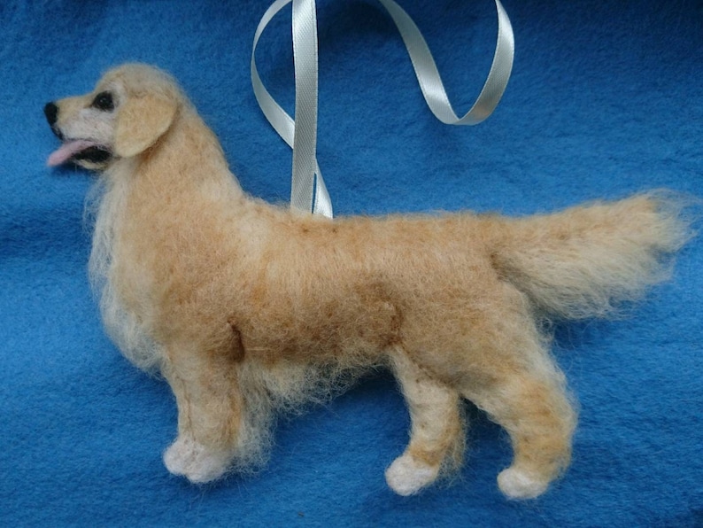 K&ouml;nnte beinhalten: Ein handgefilzter Golden Retriever Hundefiguren-Anh&auml;nger mit einem wei&szlig;en Band. Der Hund steht auf einem blauen Hintergrund.