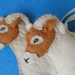 May include: Two brown and white felt horse head ornaments with stitched details. The horses are connected by a white ribbon.