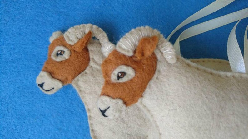 May include: Two brown and white felt horse head ornaments with stitched details. The horses are connected by a white ribbon.