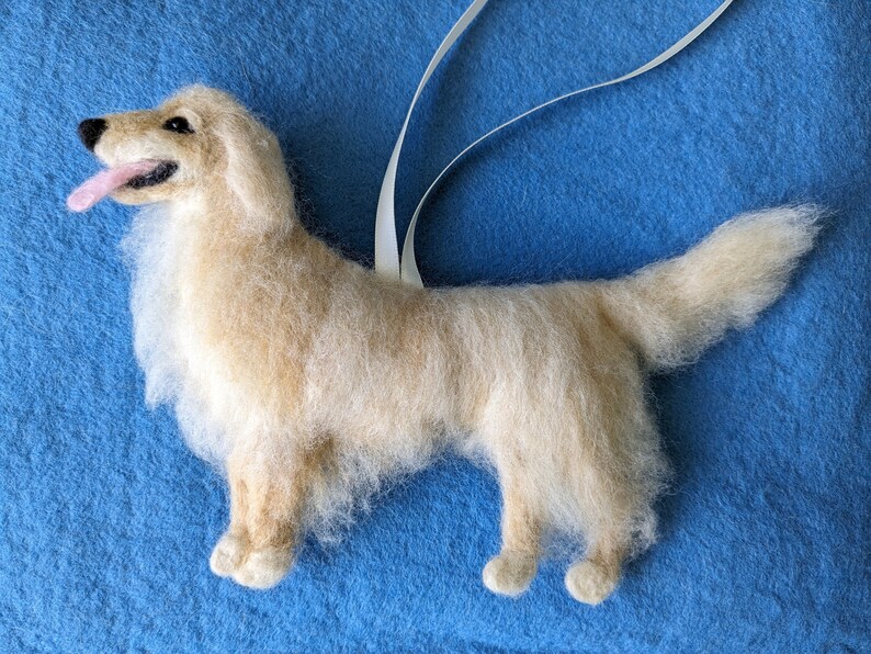 K&ouml;nnte beinhalten: Ein Nadelfilz-Anh&auml;nger in Form eines Golden Retrievers mit einem wei&szlig;en Band zum Aufh&auml;ngen. Der Hund steht auf einem blauen Hintergrund.