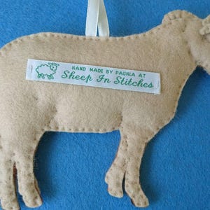 May include: A tan felt sheep ornament with a white ribbon for hanging. The sheep has a stitched-on label that reads "Hand Made by Paula at Sheep In Stitches".