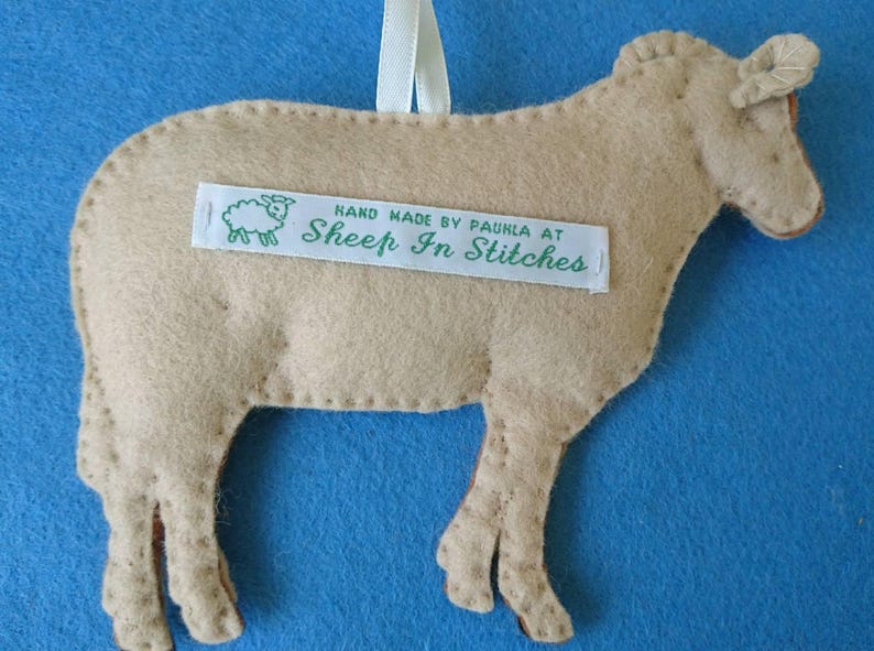 May include: A tan felt sheep ornament with a white ribbon for hanging. The sheep has a stitched-on label that reads "Hand Made by Paula at Sheep In Stitches".