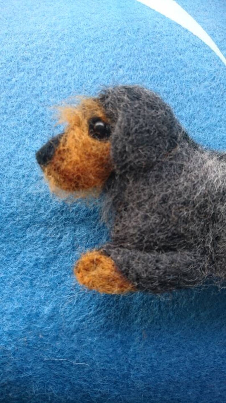 May include: A needle-felted dog figurine crafted from dark grey and tan wool fibres. The small sculpture features a textured coat, a black nose, and a dark eye, capturing the likeness of a wire-haired dachshund in a lying position.