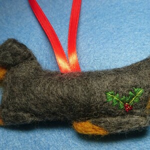 May include: A felted dog ornament in dark grey with tan accents on the face and paws. The dog features an embroidered holly sprig with green leaves and red berries on its side. A bright red ribbon loop is attached to the top for hanging.