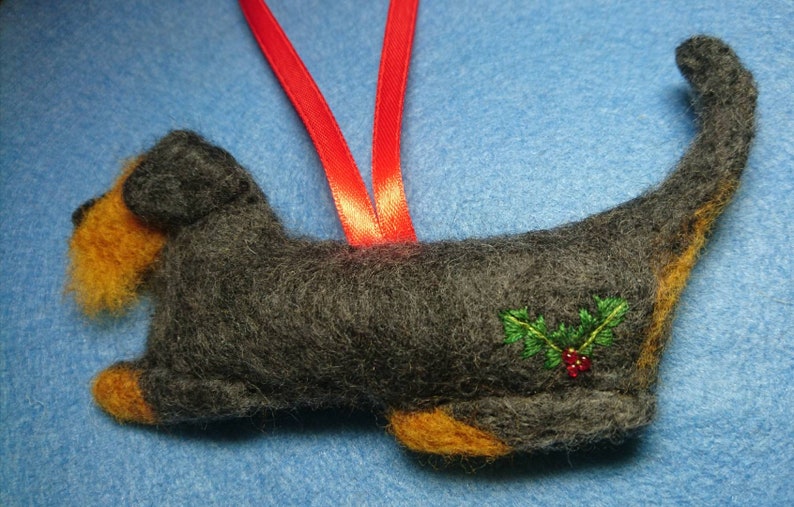 May include: A felted dog ornament in dark grey with tan accents on the face and paws. The dog features an embroidered holly sprig with green leaves and red berries on its side. A bright red ribbon loop is attached to the top for hanging.
