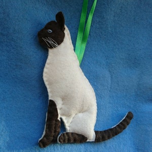 May include: A white and brown felt Siamese cat ornament with a green ribbon attached to the top. The cat is sitting with its tail curled around its legs.