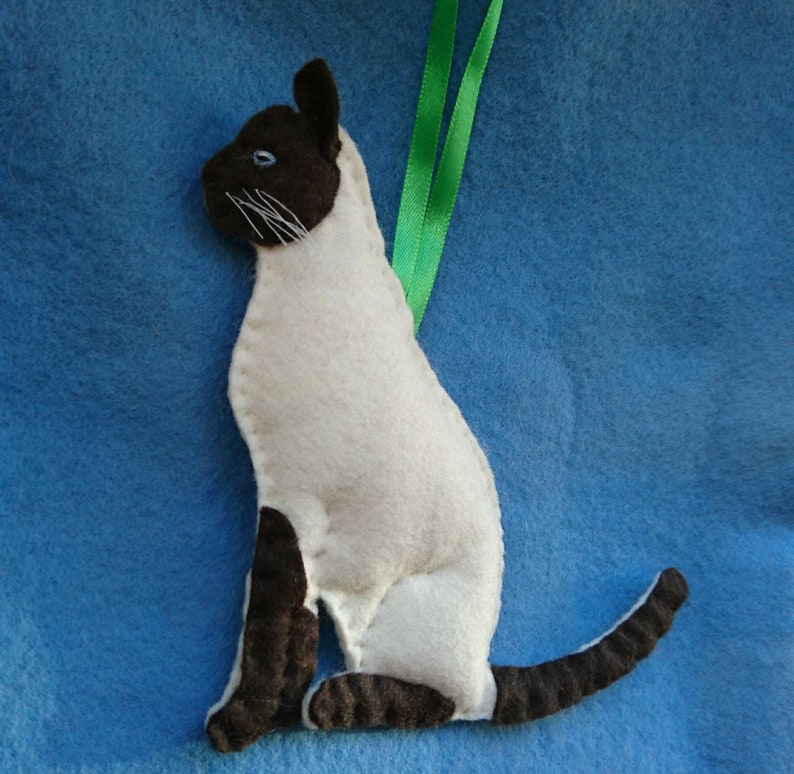 May include: A white and brown felt Siamese cat ornament with a green ribbon attached to the top. The cat is sitting with its tail curled around its legs.