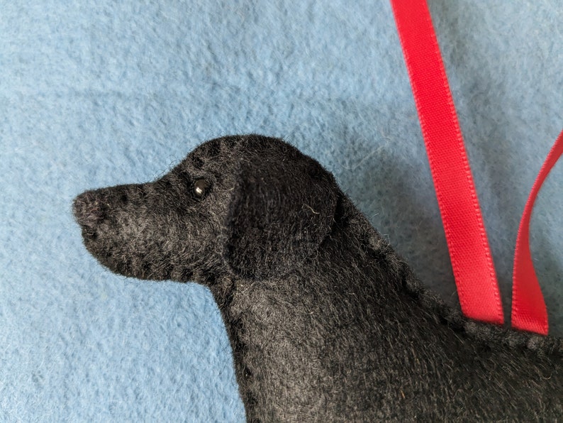 May include: Close-up of a black felt dog ornament with a red ribbon. The dog's head is turned to the side, and one eye is visible.