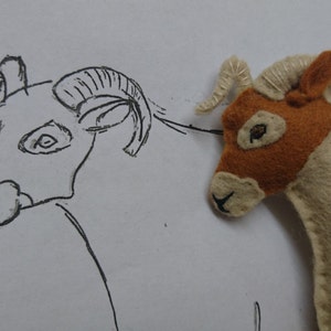 May include: A hand-stitched felt toy ram with brown and white fur, next to a pencil drawing of a ram with large horns.