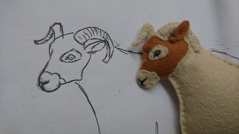 May include: A hand-stitched felt toy ram with brown and white fur, next to a pencil drawing of a ram with large horns.